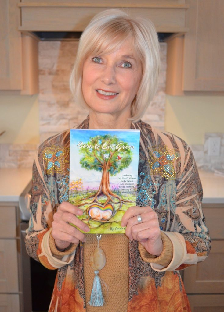 Blonde woman holding her book titled "Born to Live Green" while standing in a home kitchen.