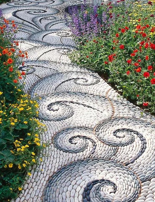 Mosaic stone pathway featuring intricate swirl patterns bordered by colorful flowering garden plants.